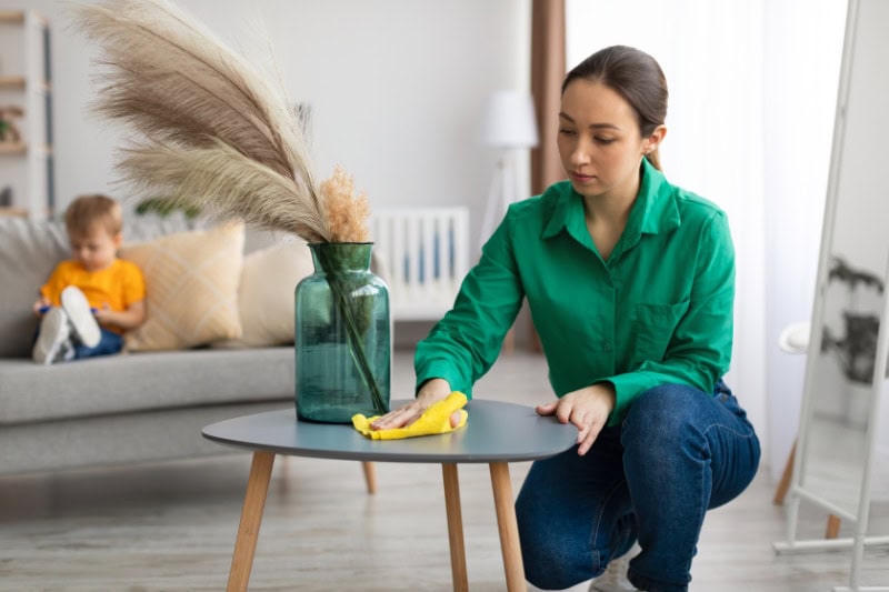 How to Reduce Dust in Your Home. Spring cleaning. Young mom dusting off coffee table and tidying up in living room while her toddler son watching cartoon on cellphone