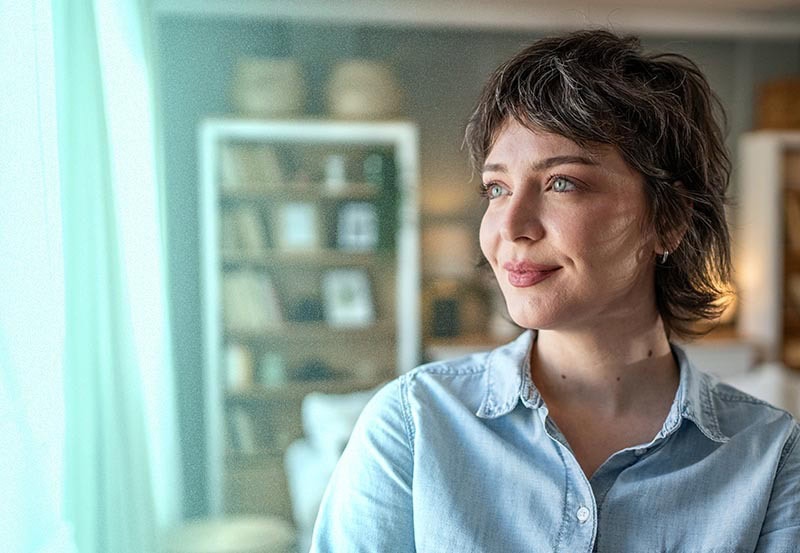 A close-up portrait of a woman indoors, looking thoughtfully toward a window.