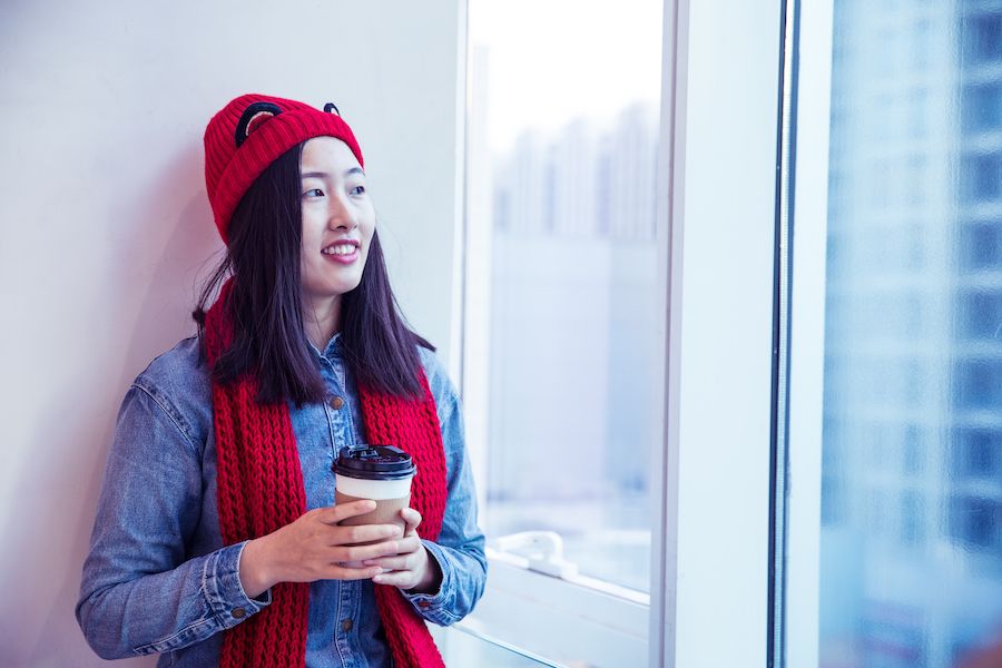The Basics of Winter Indoor Air Quality. Image shows woman inside wearing a scarf and hat while drinking coffee.